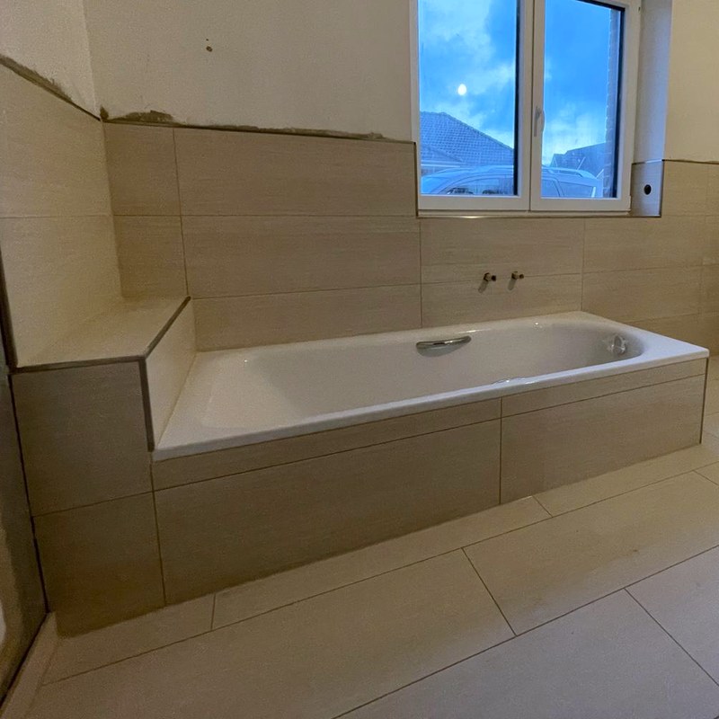 Eingeflieste Badewanne in beigen Fliesen, davor großformatige helle Bodenfliesen im Versatz